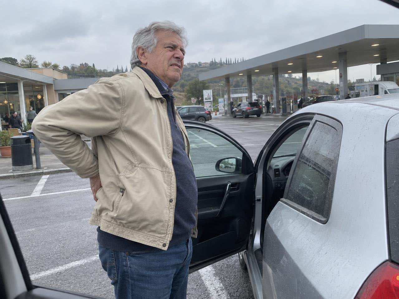Aldo in piedi accanto all'auto in un'area di servizio, con una mano sulla schiena